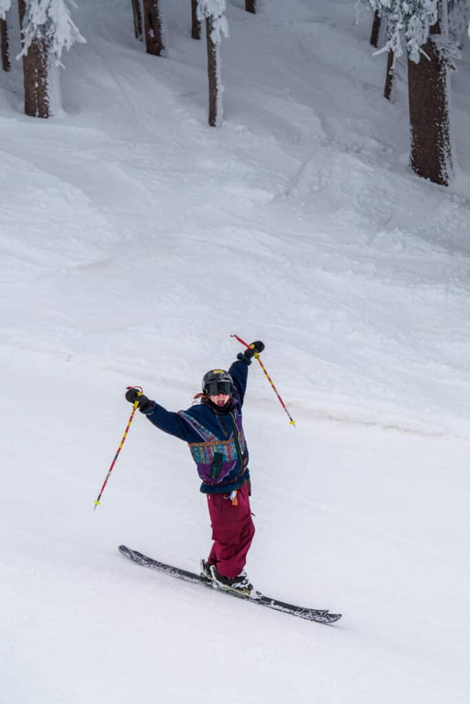 Skier wearing red pants and a colorful top holding their poles up and smiling towards the camera
